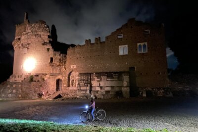 Cyclist with e‑bike at night in front of the illuminated Tomb of Cecilia Metella on the Appian Way